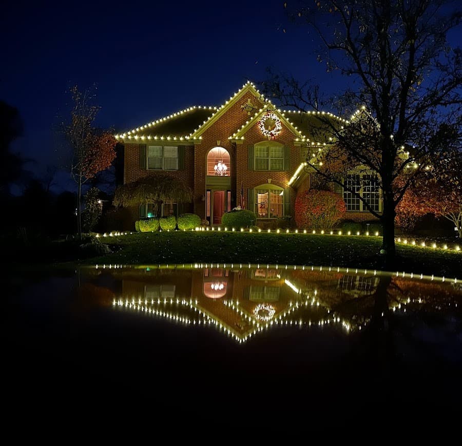 A home with warm white Christmas lights outlining the roofline, reflected in a pond at dusk